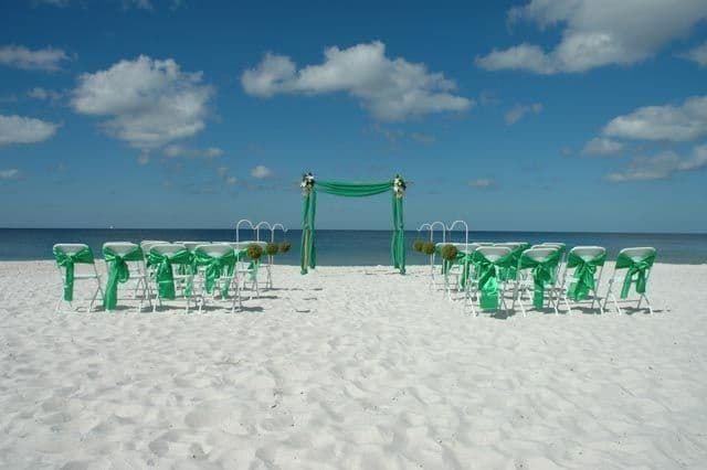 Beach wedding setup with green decorations, chairs facing an arch, against a blue sky.