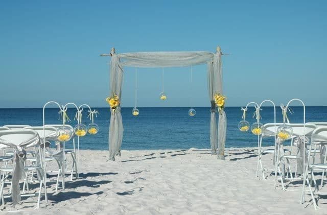 Beach wedding ceremony setup: white chairs, archway with draped fabric, ocean backdrop.