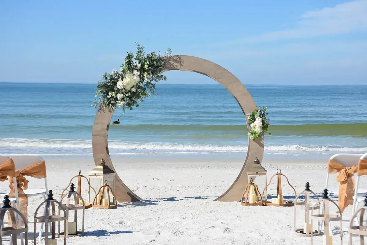 Beach wedding ceremony with floral-decorated wooden arch, white chairs, and lanterns. Ocean in background.