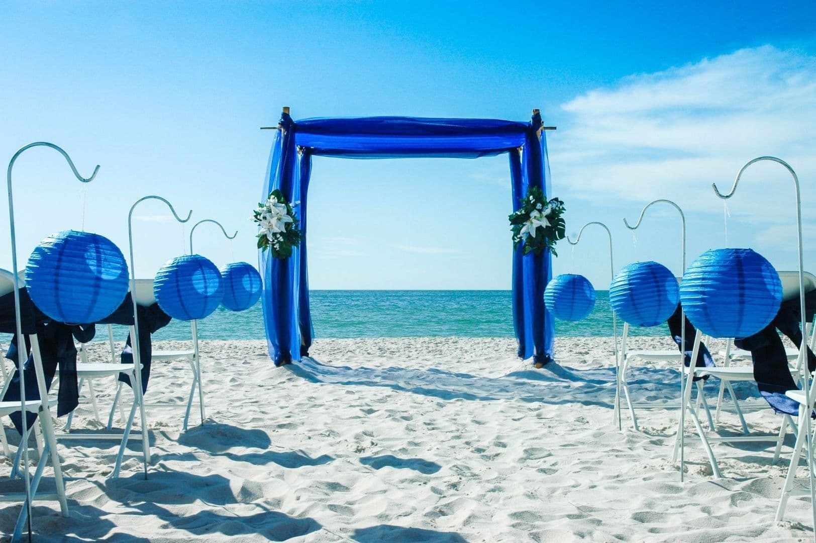 Beach wedding setup with blue arch, lanterns, and white chairs. Ocean in background.