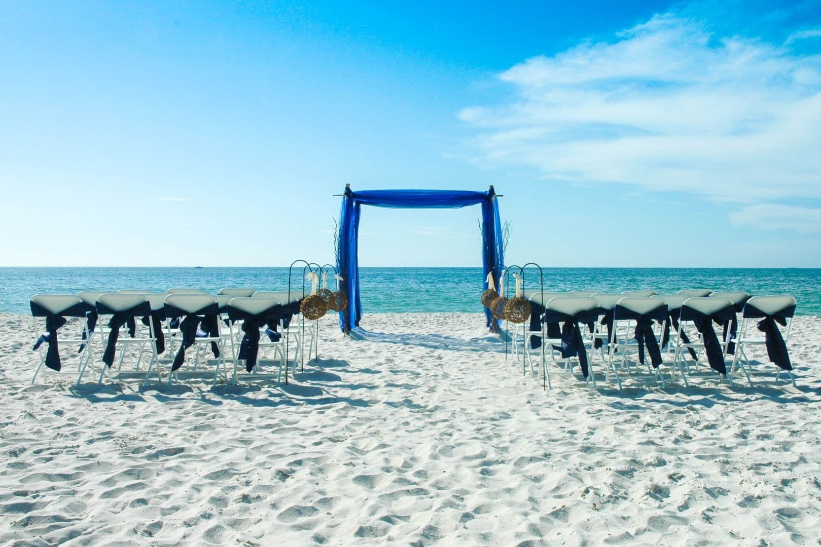 Beach wedding setup with blue arch, white sand, and chairs facing the ocean.