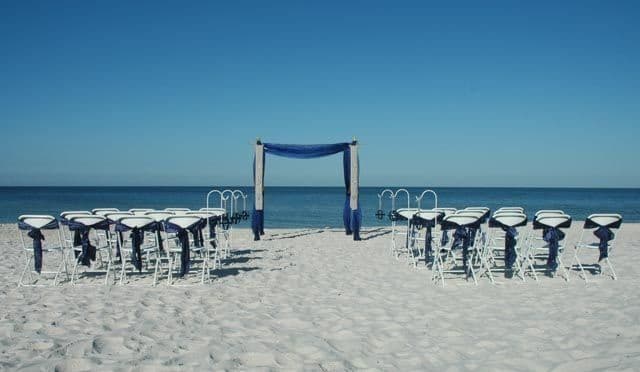 Beach wedding setup with chairs facing a draped arch, ocean backdrop.
