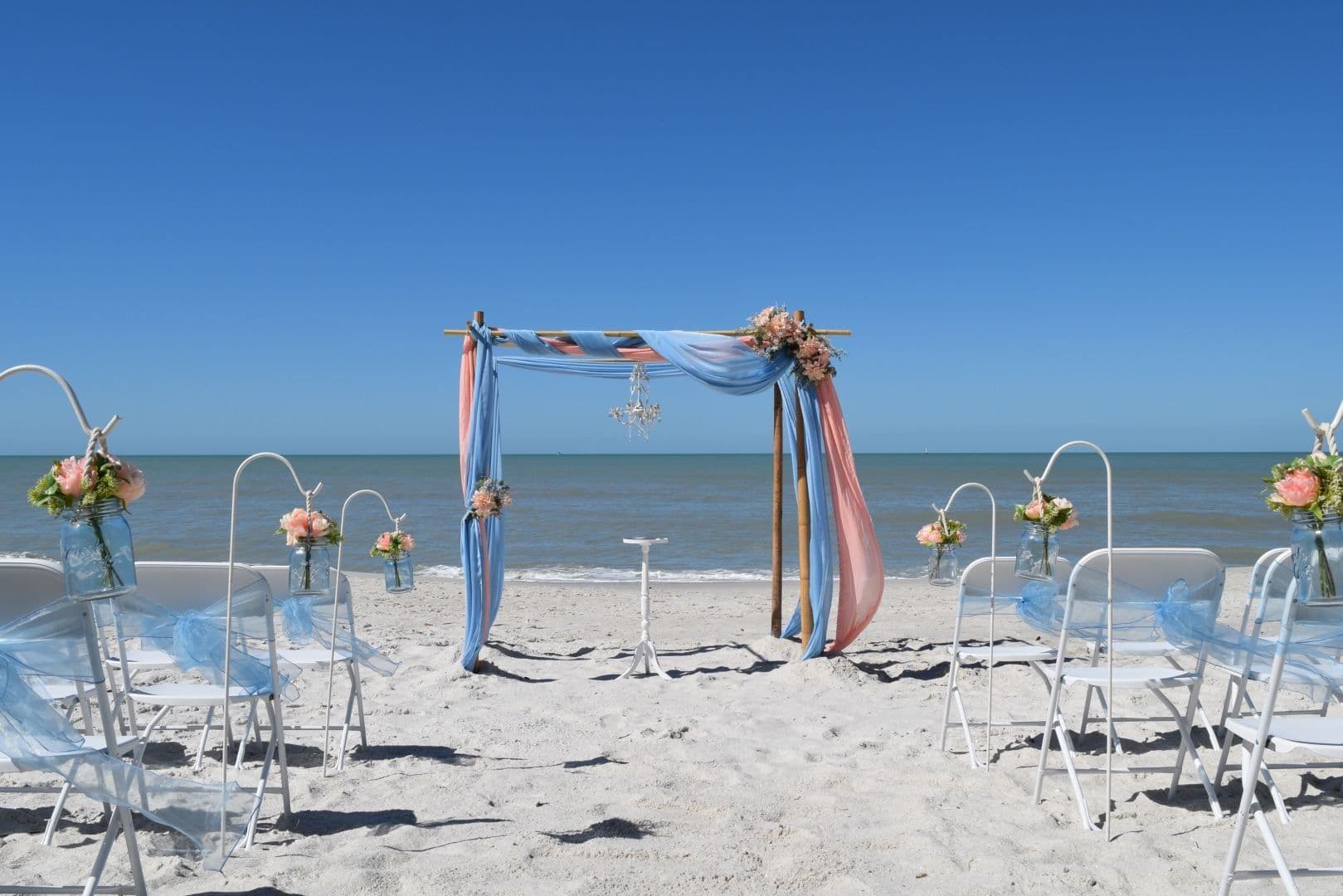 Beach wedding ceremony setup with blue and peach draped arch, chairs facing the ocean.