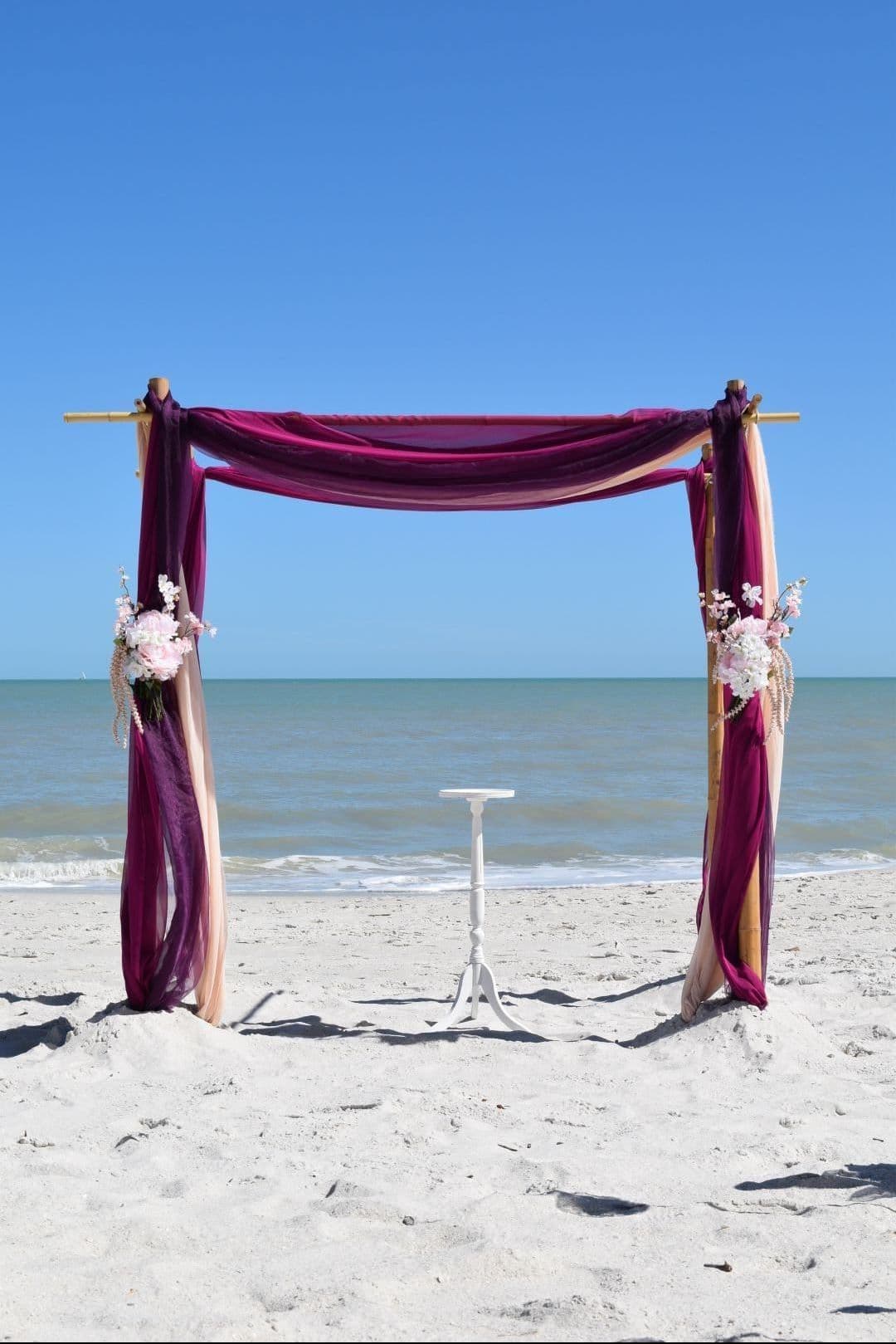 Beach wedding arch draped with purple and gold fabric, flowers, ocean backdrop.