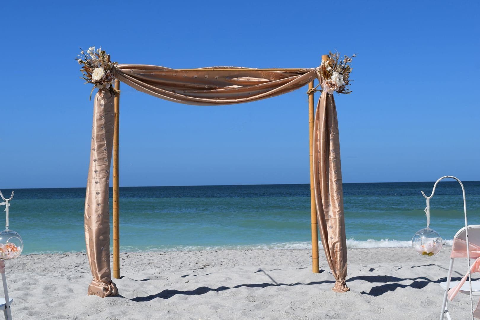 Beach wedding arch with gold fabric, flowers, and ocean background.