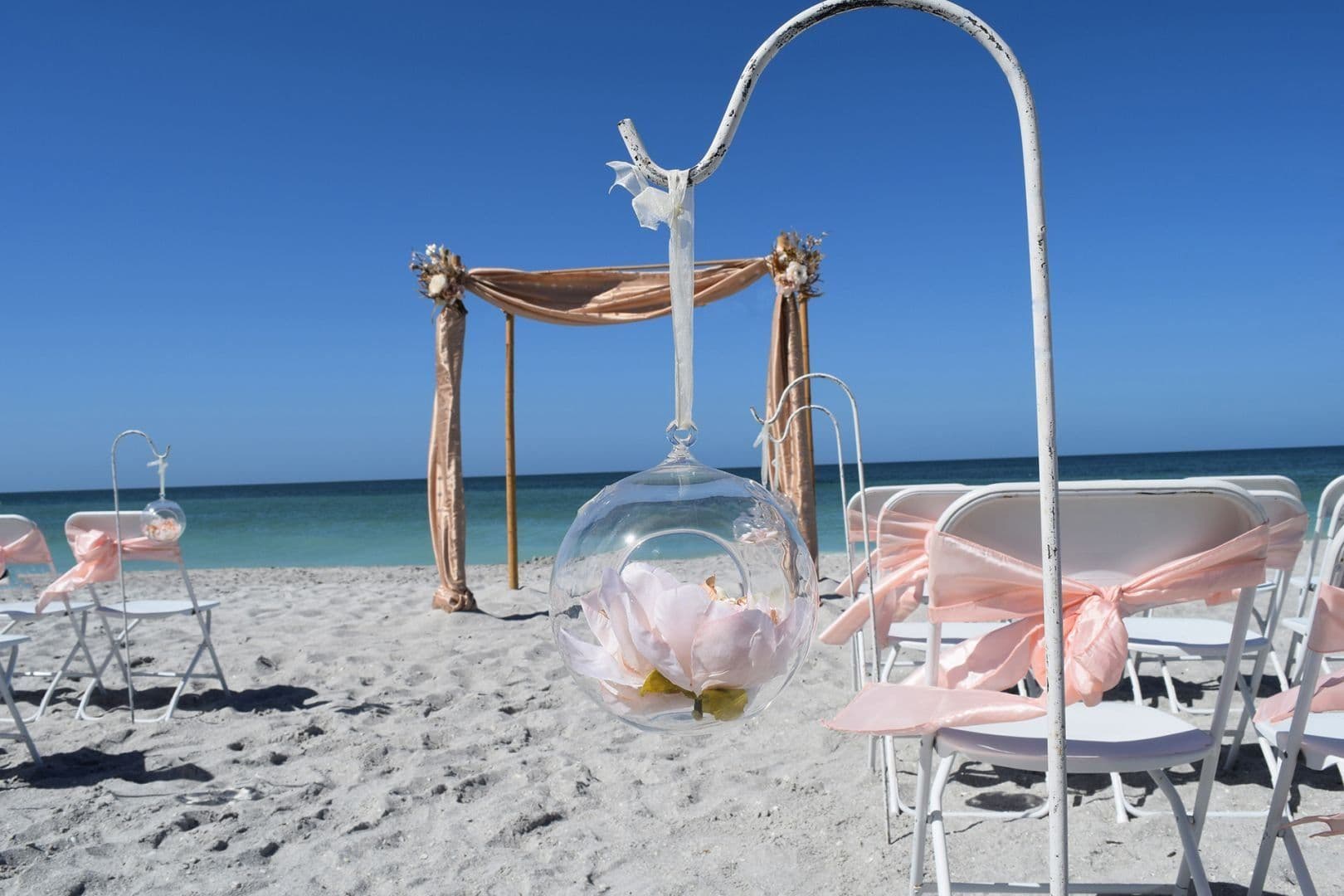 Beach wedding setup: white chairs with pink bows, floral arch, clear globe with flowers, blue sky and ocean.