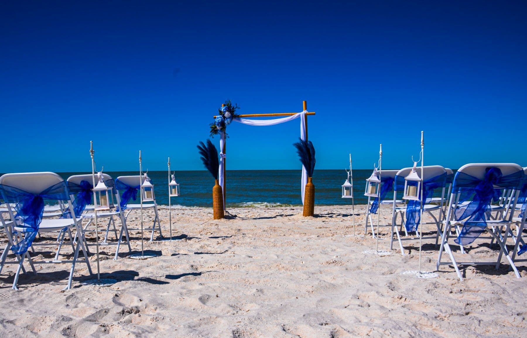 Beach wedding setup: chairs with blue sashes face an archway with ocean backdrop.