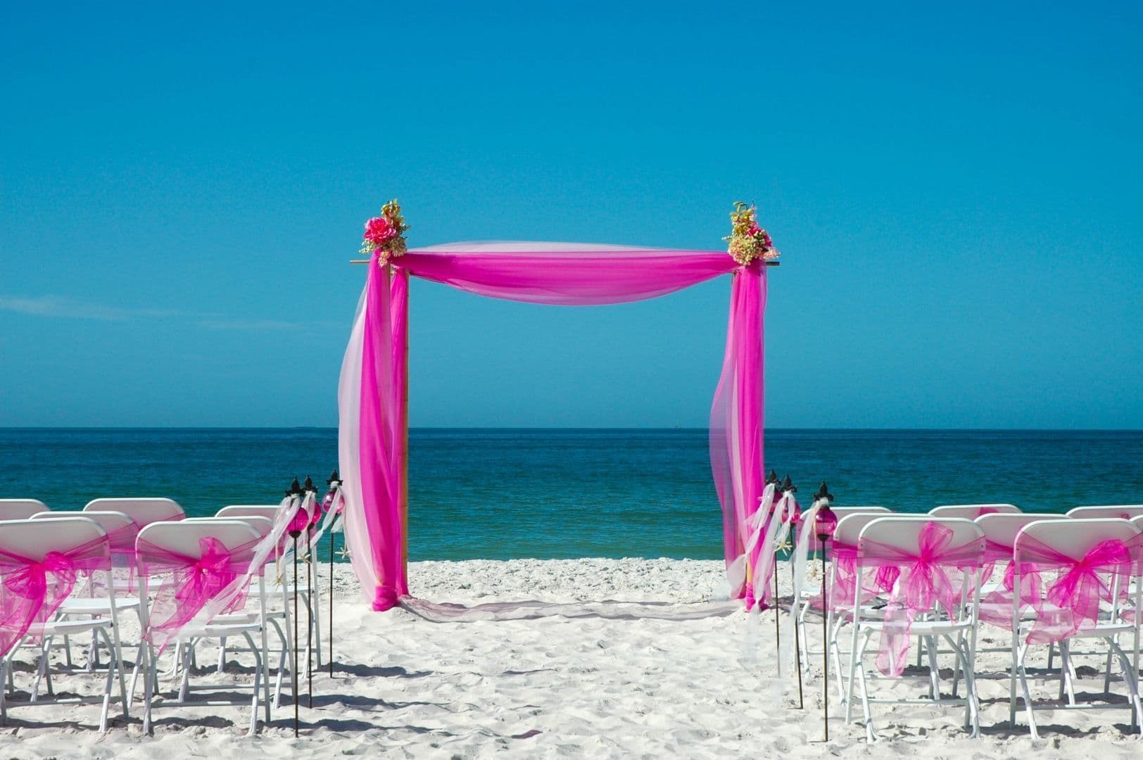 Beach wedding setup with pink draped arch, white chairs, and ocean backdrop.