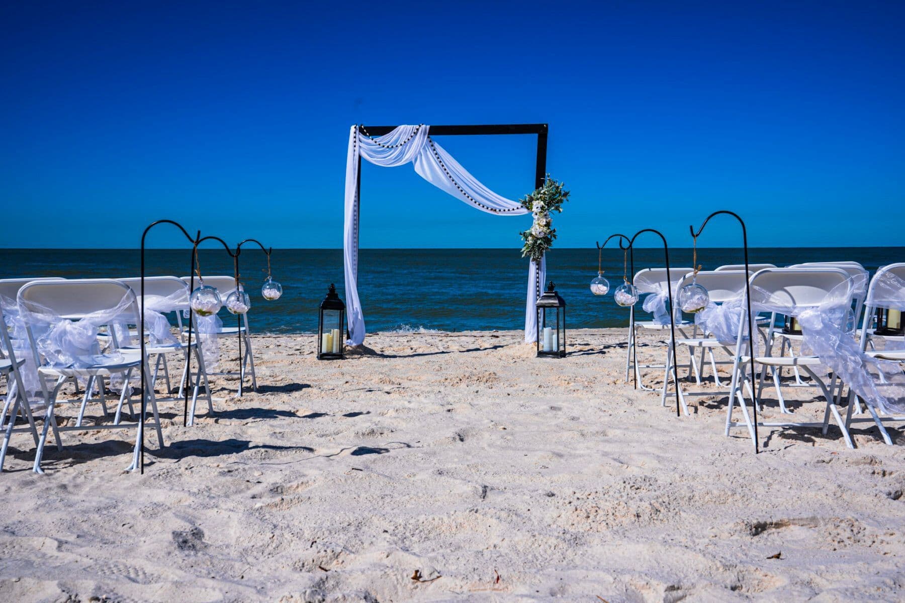 Beach wedding setup with white chairs, arch, and ocean backdrop.