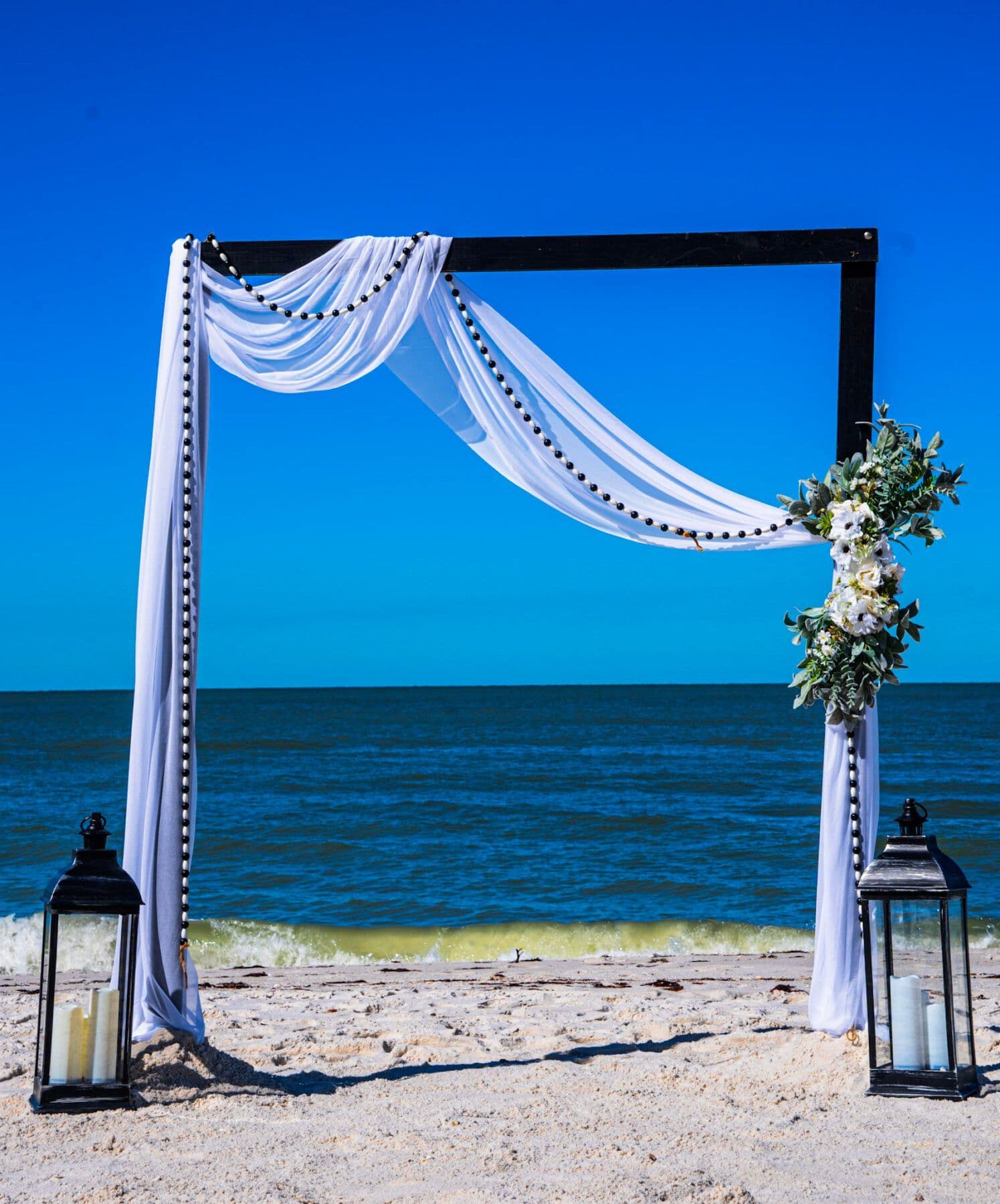 Wedding arch on a beach: black frame, white fabric and flowers, against a blue sea and sky.