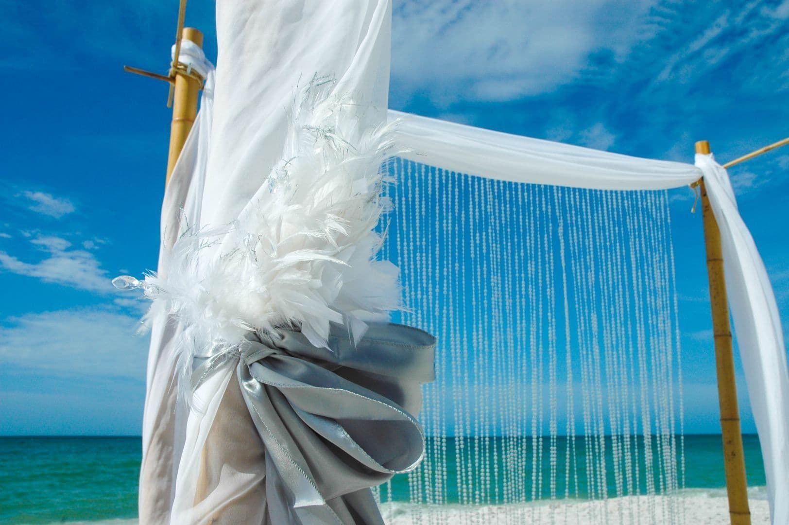 Beach wedding arch with white fabric, beads, and bamboo against a blue sky and ocean.