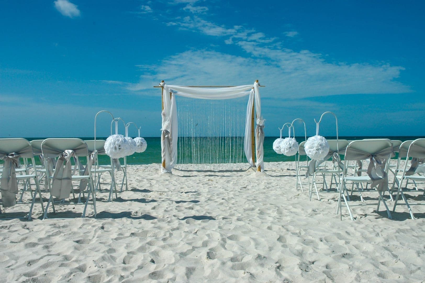 Beach wedding ceremony setup: white chairs, arch, and decorations on the sand with a blue ocean and sky background.