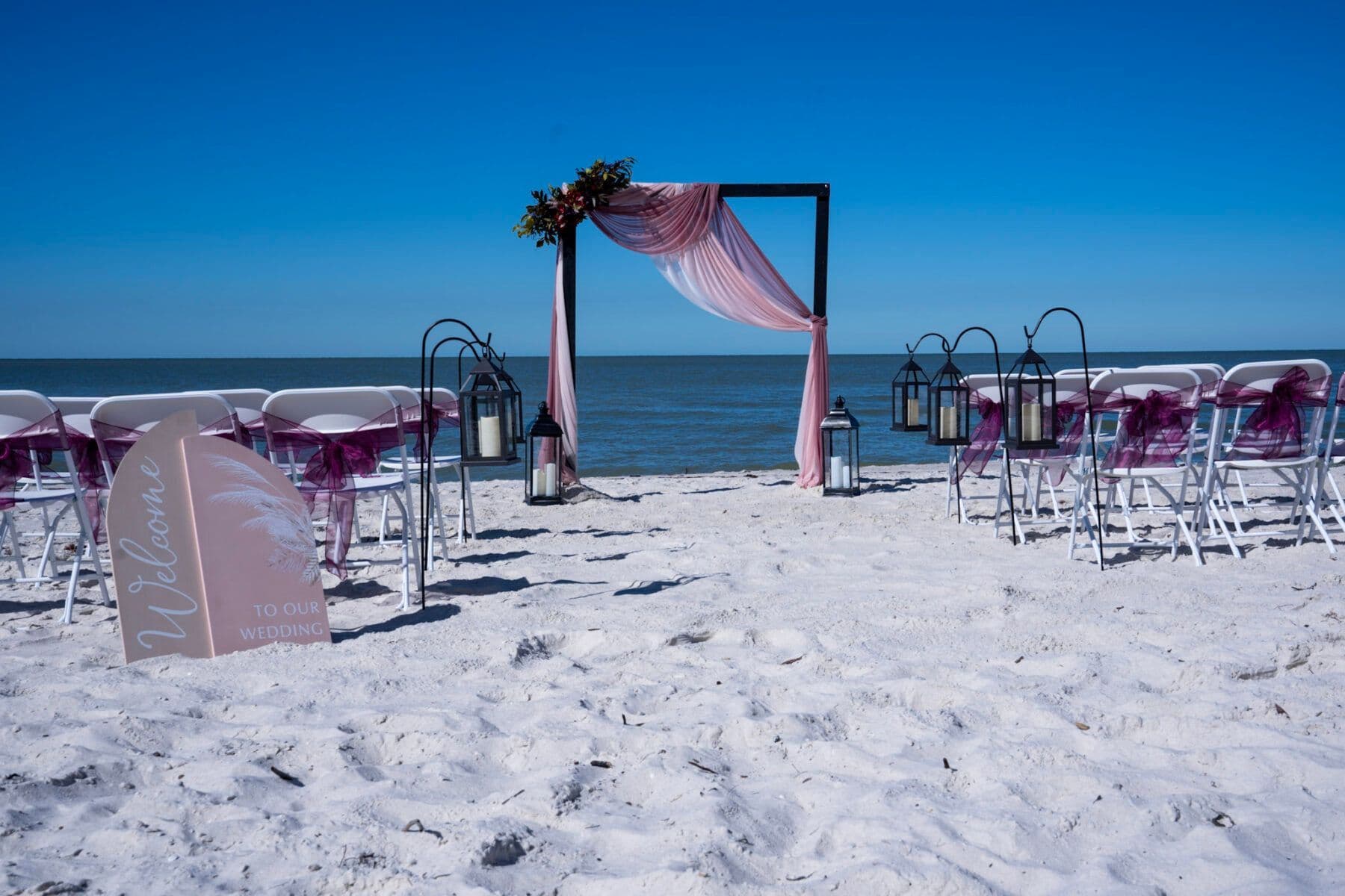 Beach wedding setup: white chairs, arch draped with pink fabric, and lanterns on the white sand with ocean backdrop.