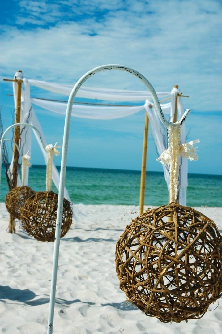 Beach wedding ceremony, white sand, clear blue sky, draped white fabric, wicker spheres, light blue water.