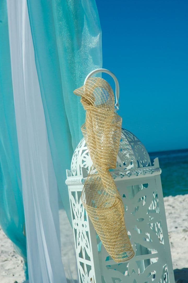 White decorative lantern with woven gold ribbon on a beach; light blue and white fabric in background.