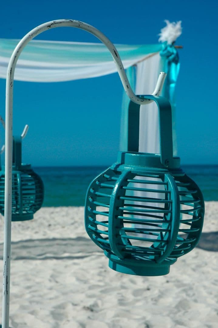 Teal lanterns hanging on white poles at a beach wedding, with ocean and blue sky backdrop.