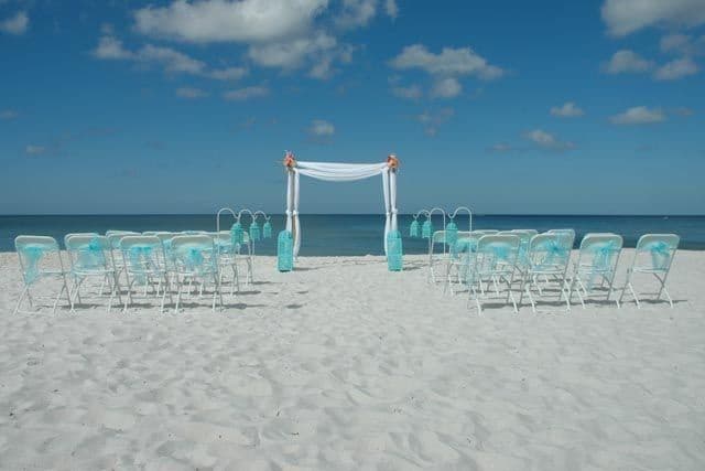 Beach wedding setup: white chairs, arch draped in white fabric, turquoise accents, ocean backdrop.