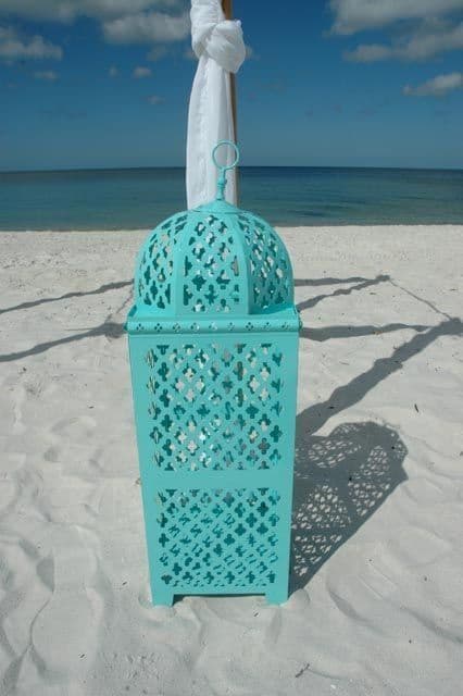 Turquoise lantern on a white sandy beach, under a wedding arch, blue ocean in the background.