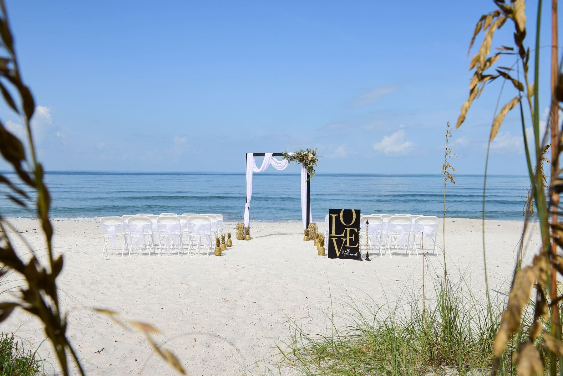 Beach wedding setup: white chairs, arch, and sign on sand with ocean backdrop.