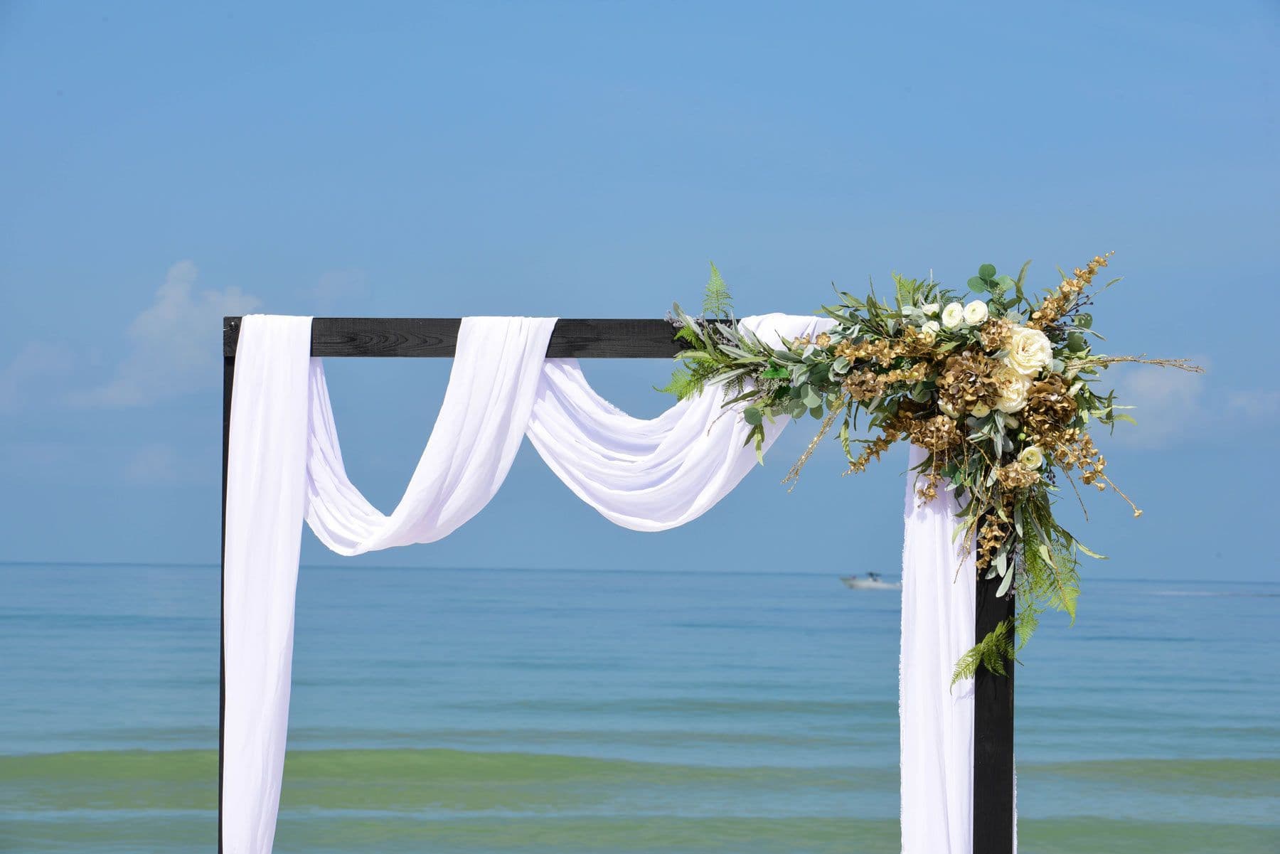 Wedding arch on beach with white fabric and floral arrangement, ocean background.