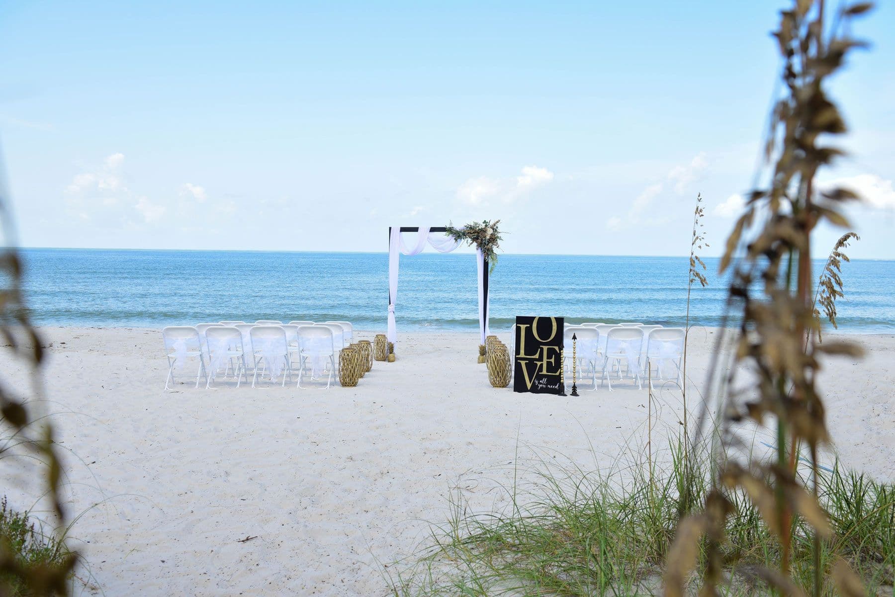 Beach wedding ceremony setup: white arch, clear chairs,