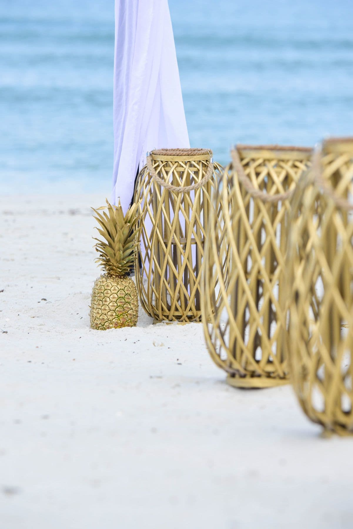Beach scene with gold lanterns and a pineapple; white fabric backdrop, ocean in background.