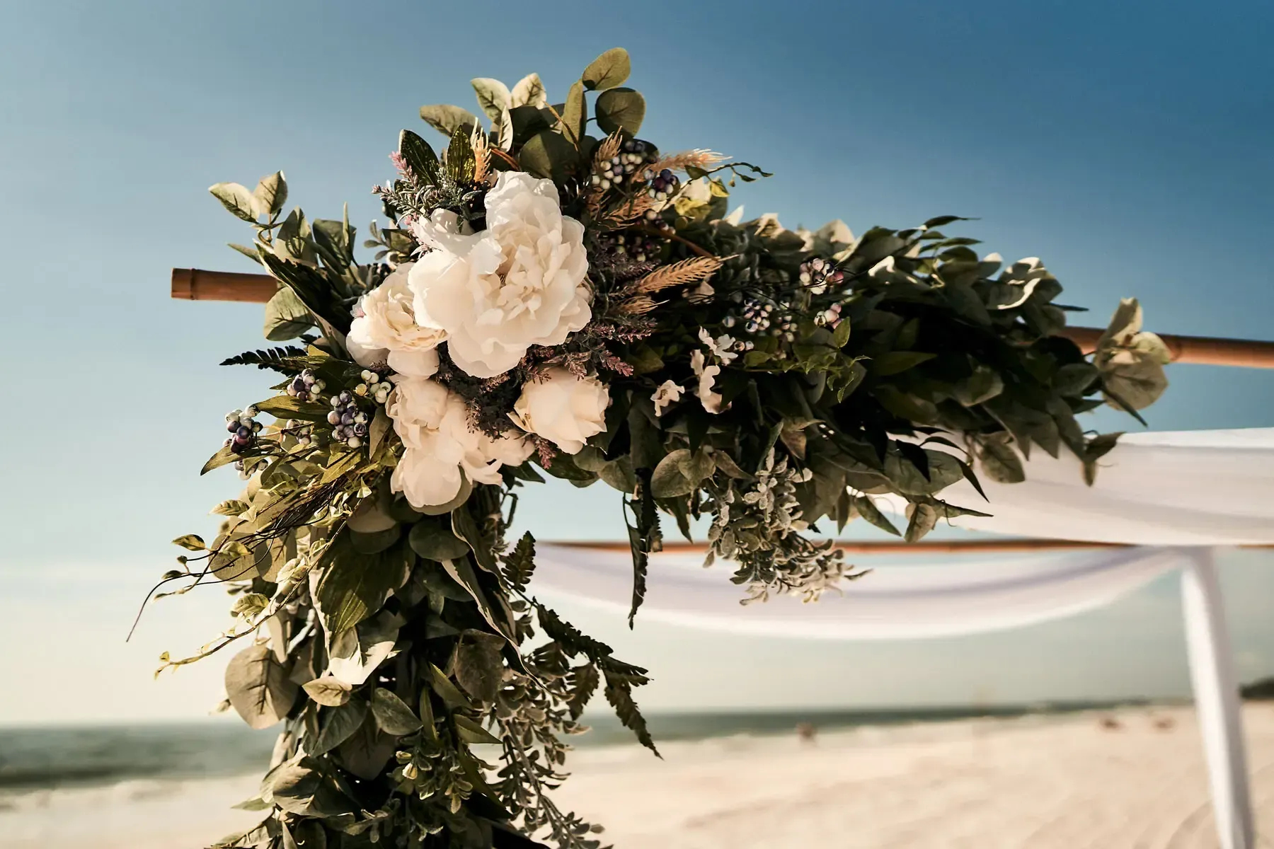 Floral arch decorated with white flowers and greenery on a beach with blue sky in background.
