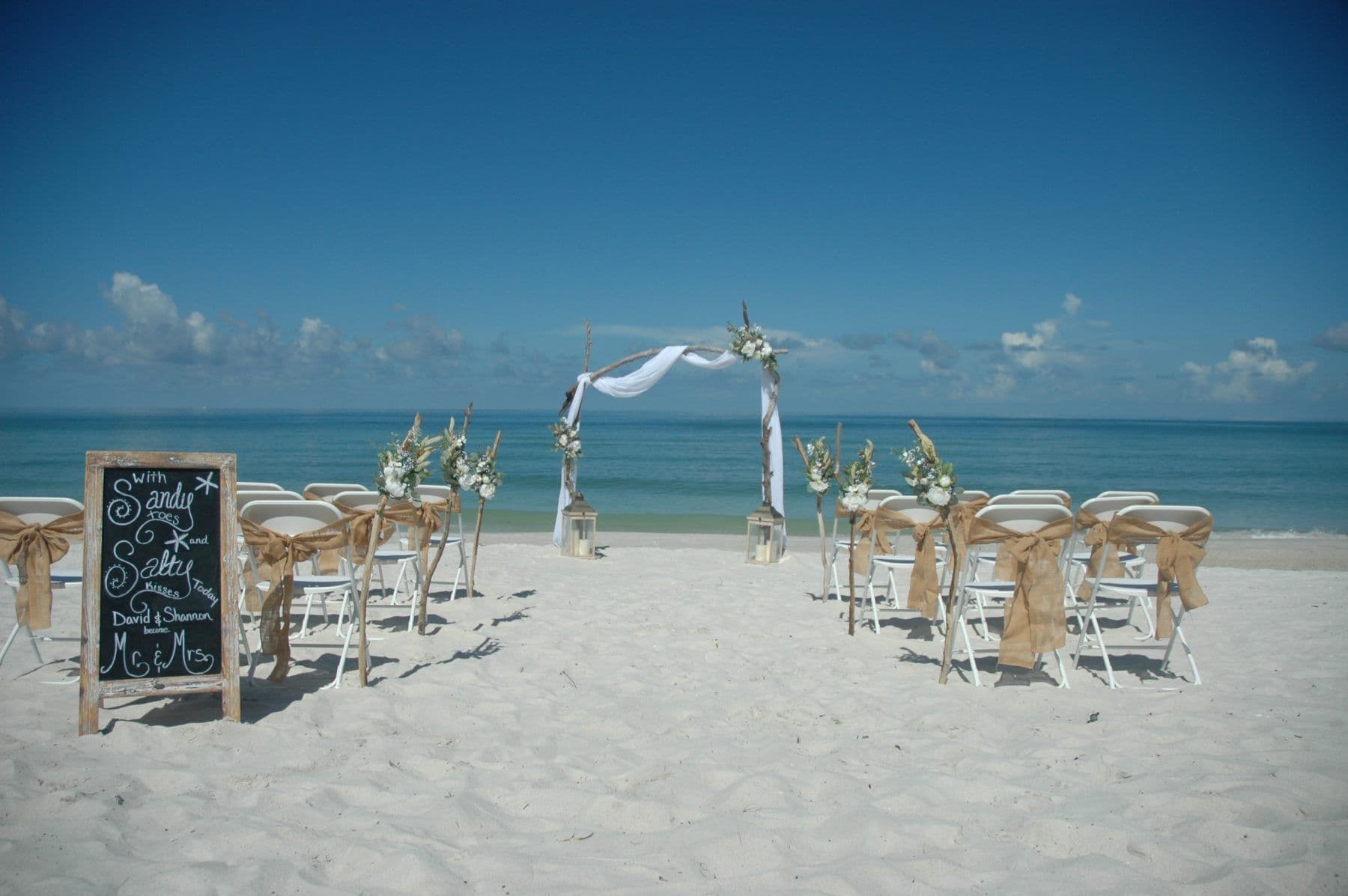 Beach wedding ceremony setup: white chairs, burlap, arch with flowers, chalkboard sign, blue sky, and ocean.