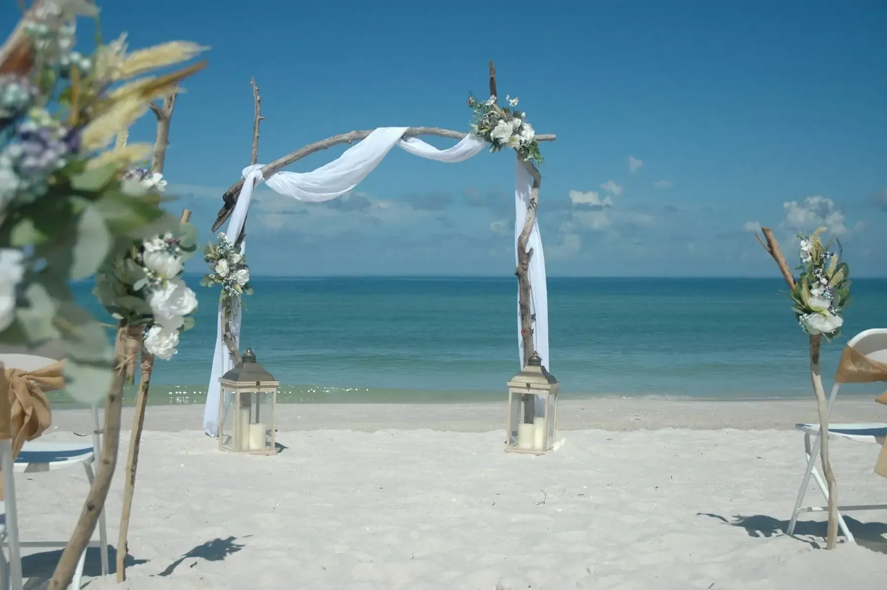 Beach wedding arch with white fabric, flowers, and lanterns, overlooking the ocean.