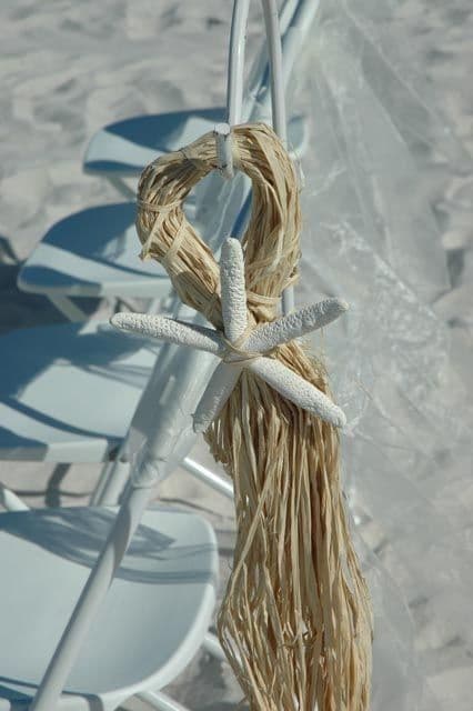 White chair with tied raffia and a starfish decoration, on a sandy beach.