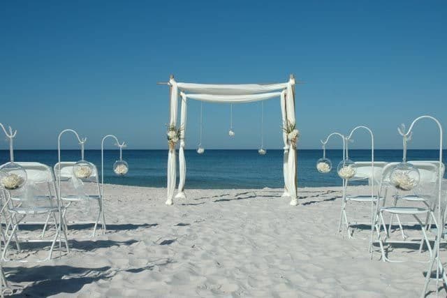 Wedding ceremony setup on a white sandy beach with blue sky and ocean. White chairs face a draped archway.