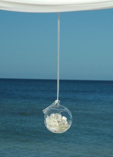 Clear glass orb with white flowers hanging from white fabric over ocean.
