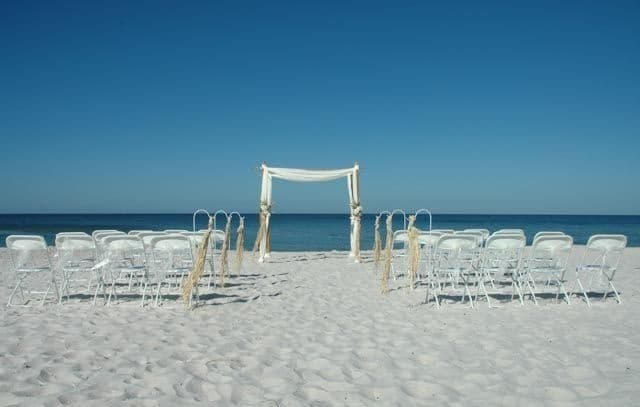 Beach wedding setup: white chairs, arch, ocean, and clear blue sky.