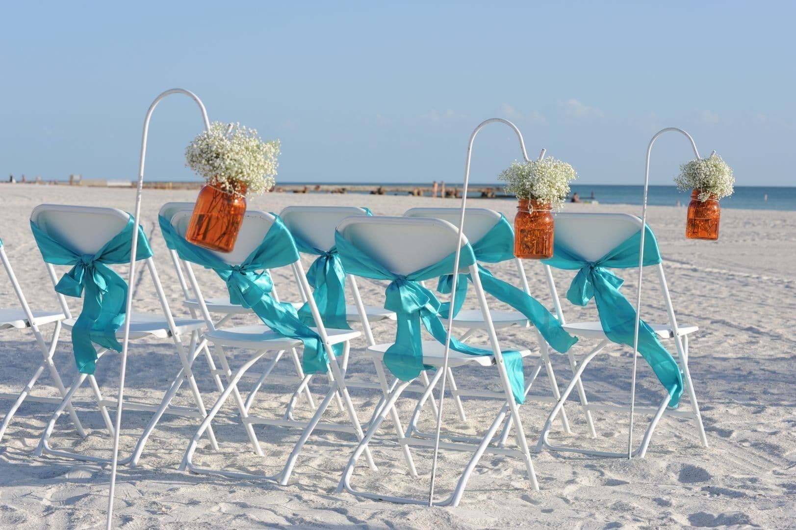 White chairs with teal bows and floral arrangements on a sunny beach, set for a ceremony.