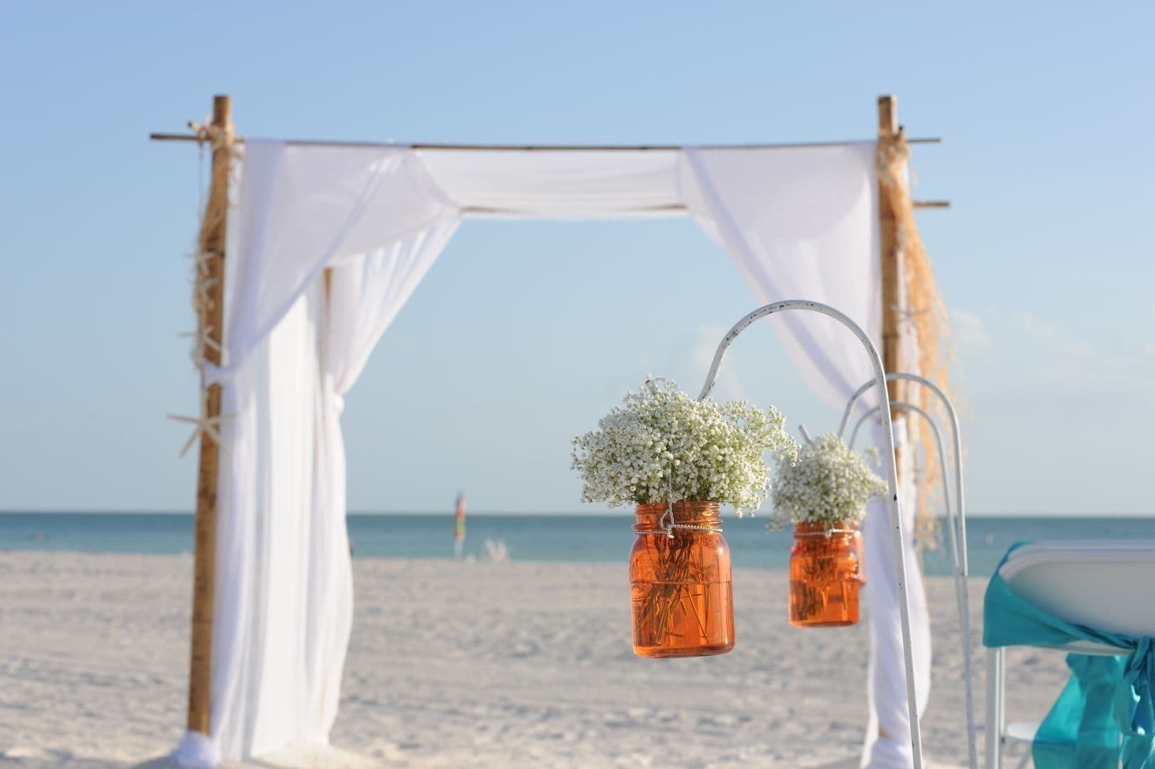 Beach wedding arch decorated with white fabric and orange flower jars.