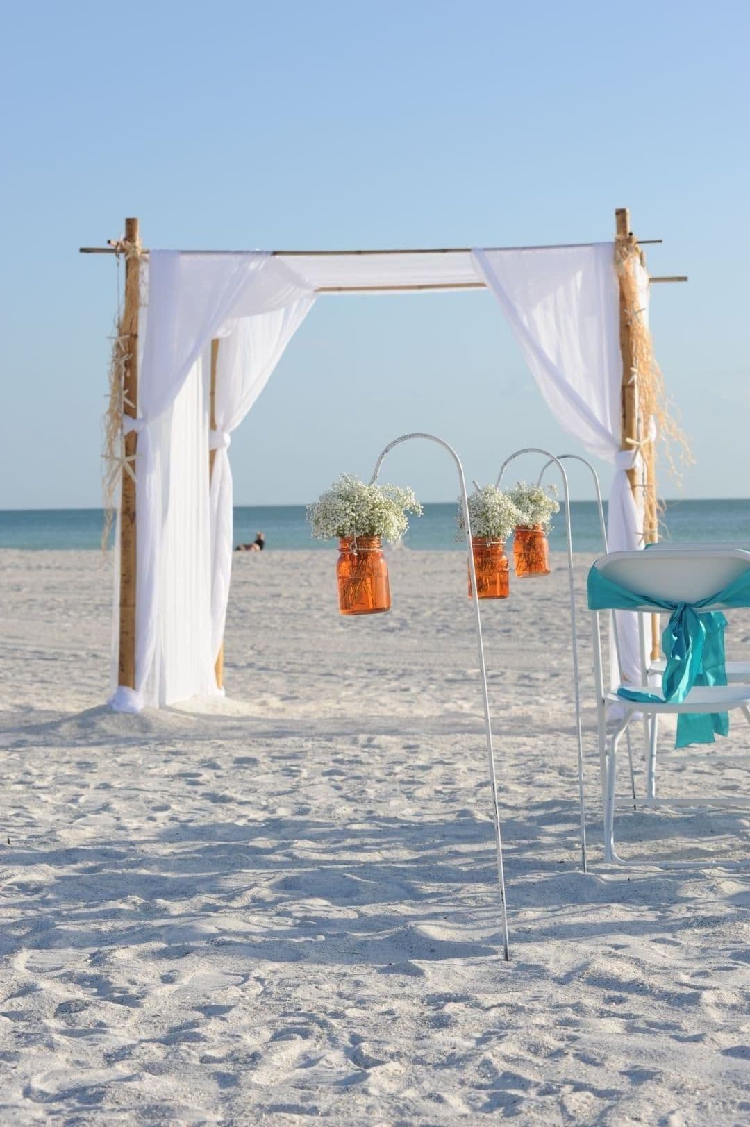 Beach wedding ceremony arch with white fabric, orange jars of flowers, and chairs on sand.